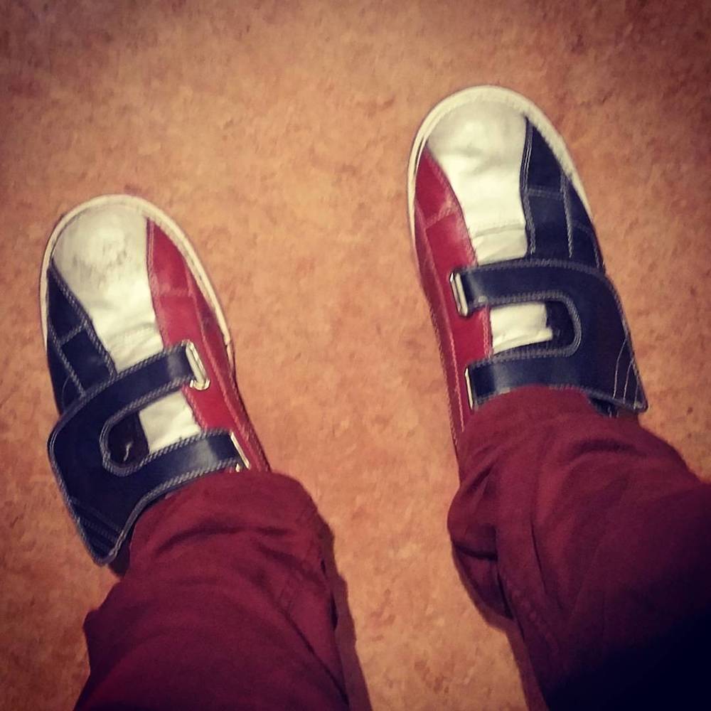 Looking down at red and blue sneakers on a patterned floor, wearing dark red trousers.