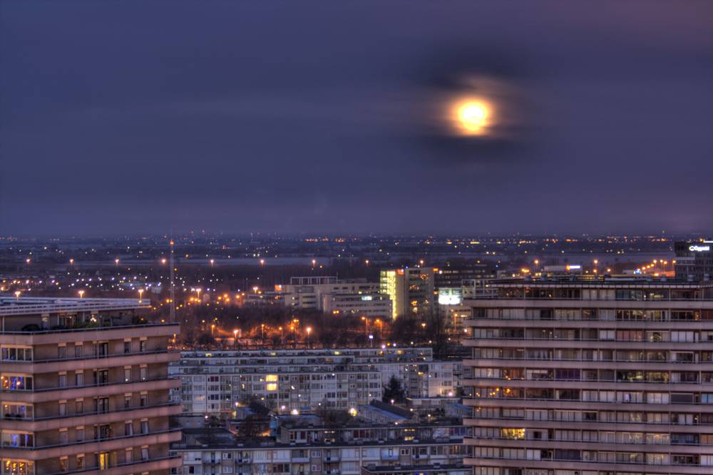 The supermoon fades behind hazy clouds above a city skyline at dawn.