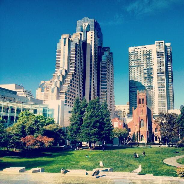 City skyline with modern towers, a red-brick church, and a park with a lawn.