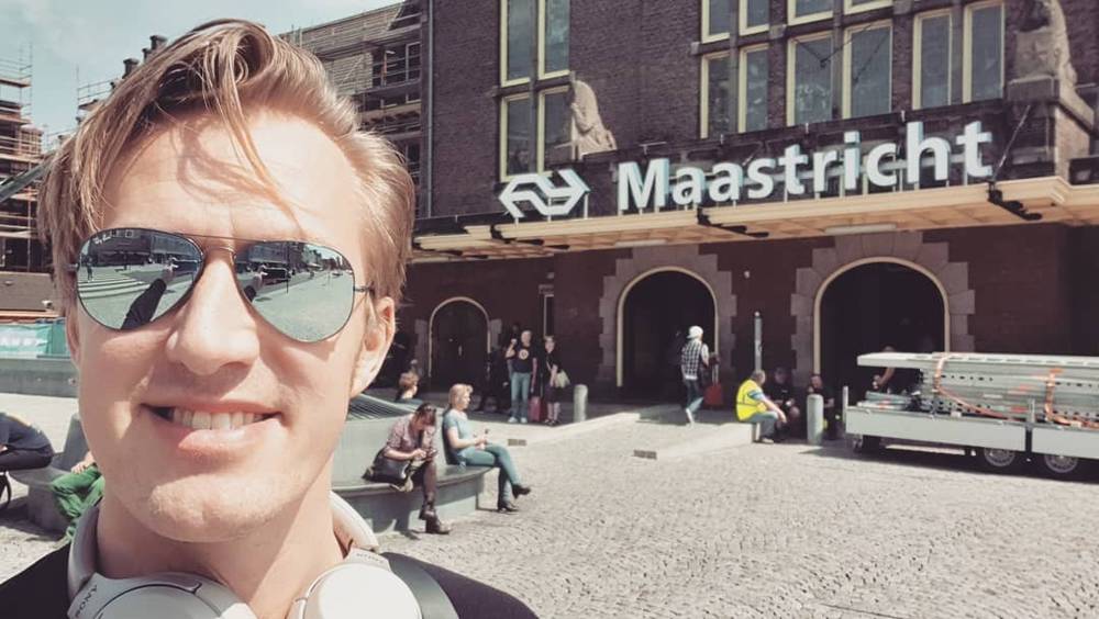 A man wearing sunglasses and headphones takes a selfie in front of the Maastricht train station sign.