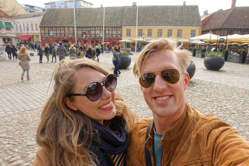 A couple smiles for a selfie in a busy town square with historic buildings.