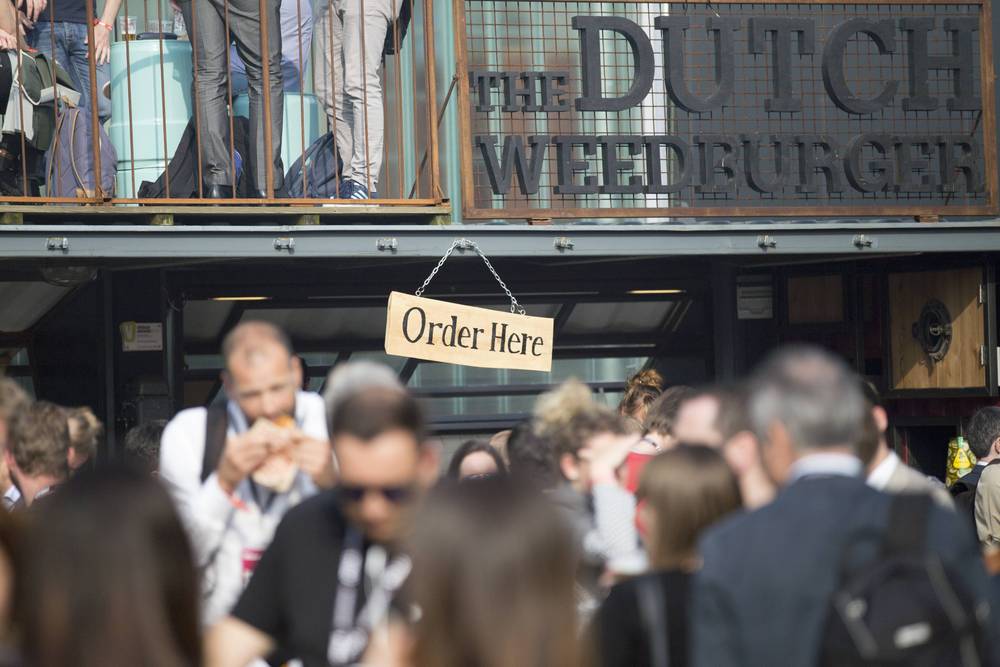A crowd eats and queues beneath 'The Dutch Weedburger' signage at a festival food stand.