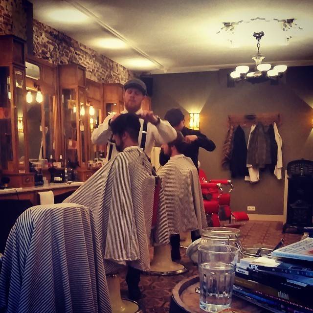 Men getting haircuts at barber chairs inside a traditional barbershop with wood paneling.