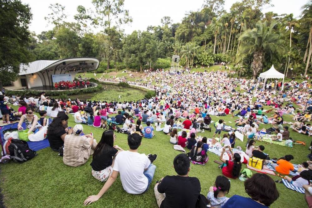 A large crowd sitting on the grass at an outdoor concert in a park.