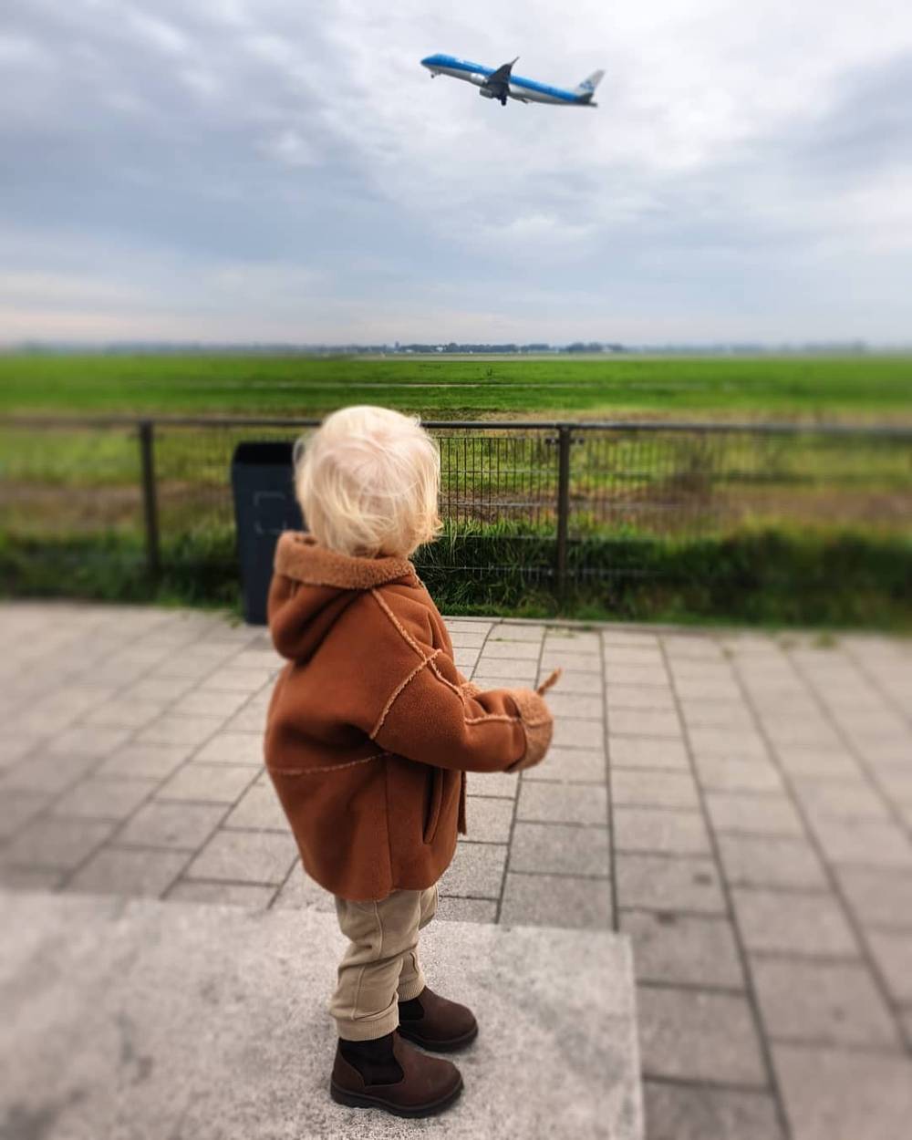 A child in a brown coat looking up at a passing airplane over open green fields.