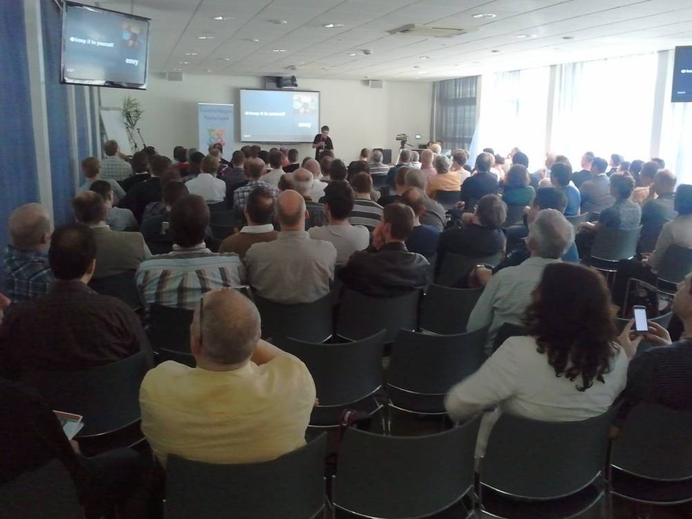 An audience watches a speaker present at the front of a conference room.