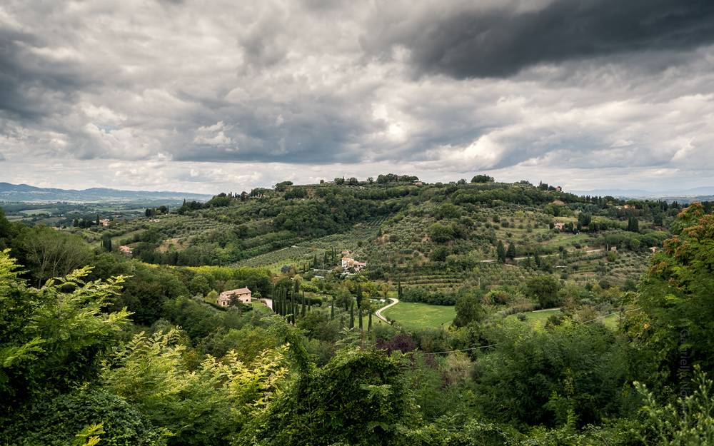 Rolling hillside covered in olive groves and cypress trees under a dramatic, heavily clouded sky, with a stone farmhouse nestled in the valley below and a hilltop village visible in the distance.