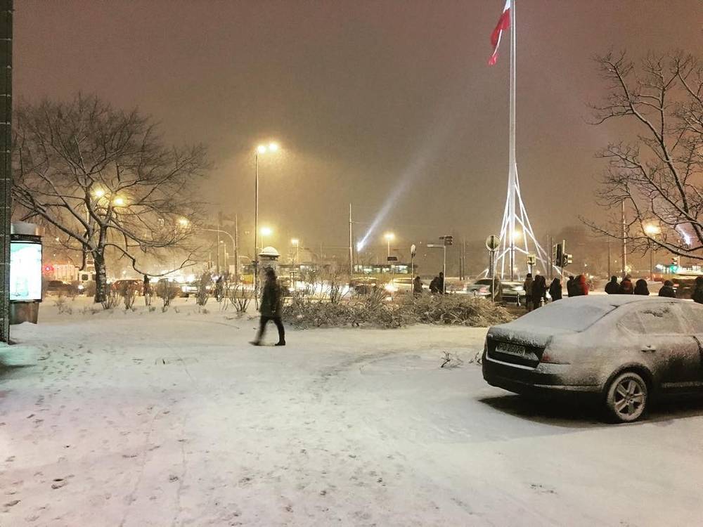 Pedestrians crossing a snowy plaza at night near a decorative light pole.