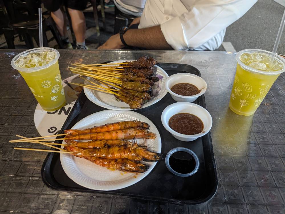 A meal on a table showing dishes of satay, shrimp and cups of sugar cane juice 