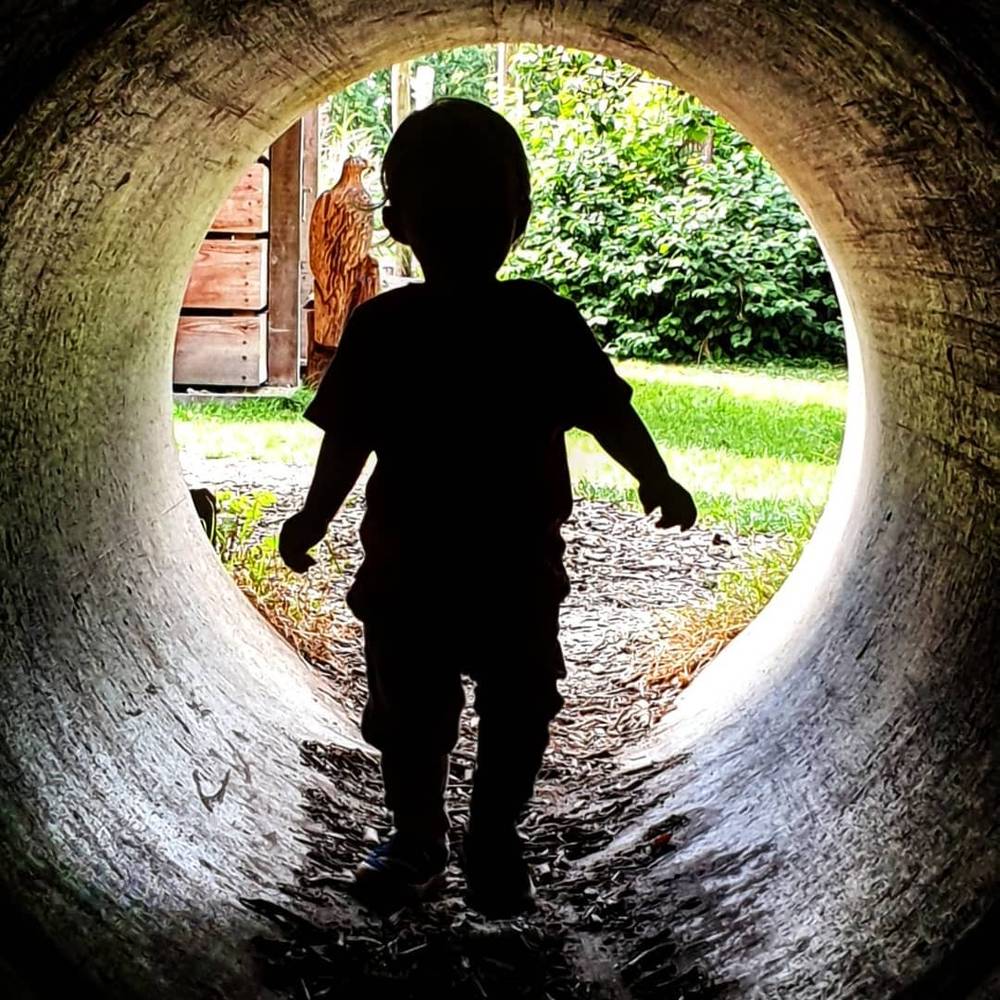 A toddler's silhouette walking through a round tunnel, backlit by greenery outside.