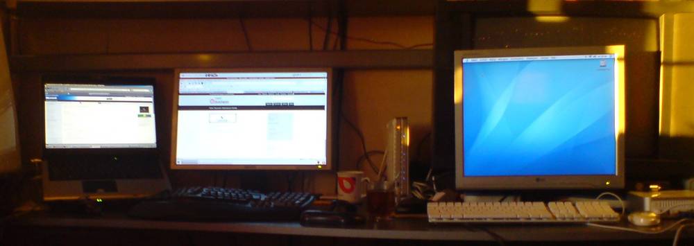 A cluttered desk with three monitors, a coffee mug and a thermometer, lit only by the screens.