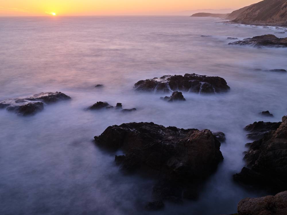 Long exposure at sunset, the sun just touching the horizon under an orange sky. Smooth misty water flows around dark rock outcroppings in the foreground, with coastal headlands fading into the distance.
