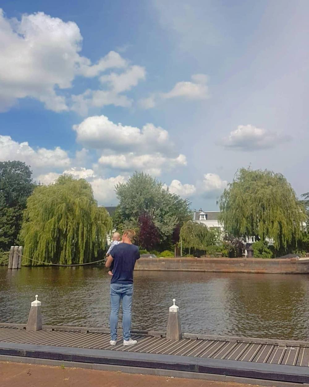 A man stands on a wooden dock by a canal at dusk, willow trees overhead.