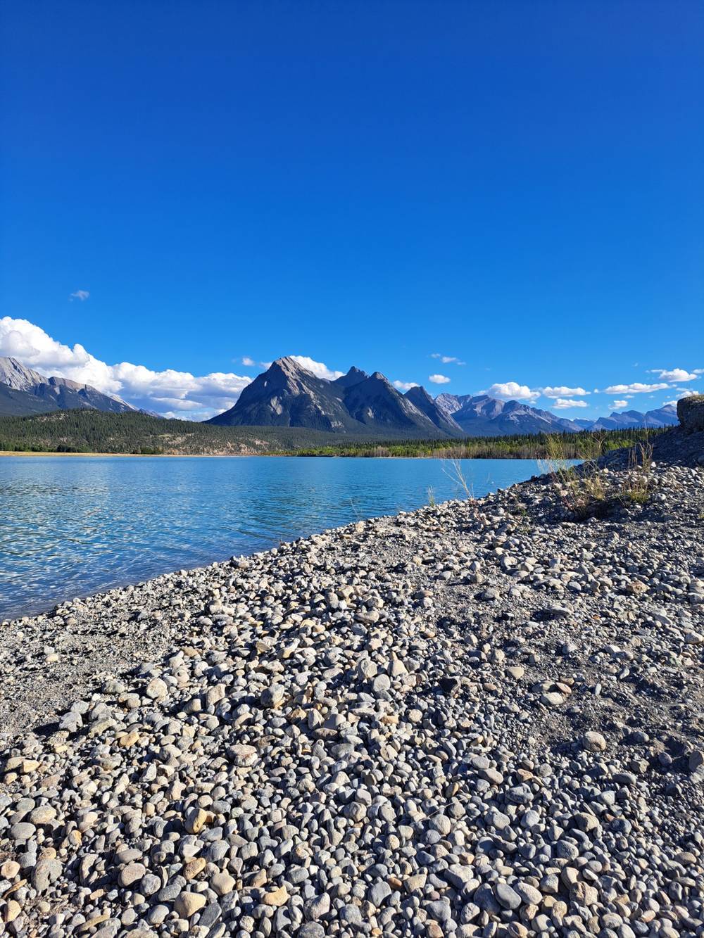 Taken from the rocky shore looking out over the lake towards the mountains on a sunny day with a blue sky and white fluffy clouds.