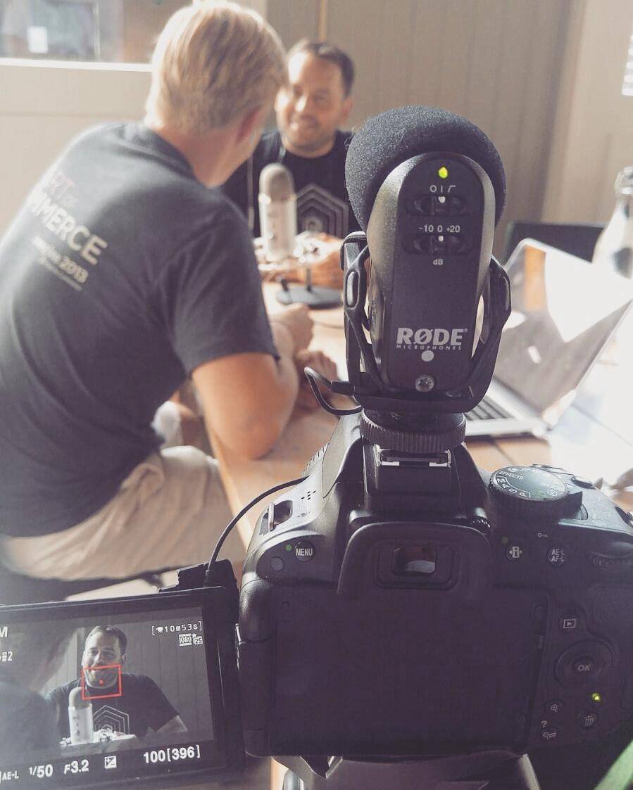Two men recording a podcast, one holding a large microphone, seen through a camera viewfinder.