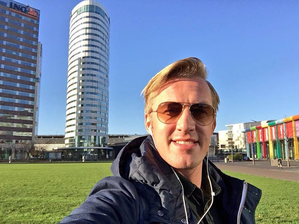 A selfie of a smiling man in sunglasses in front of a tall glass office tower and colorful buildings.