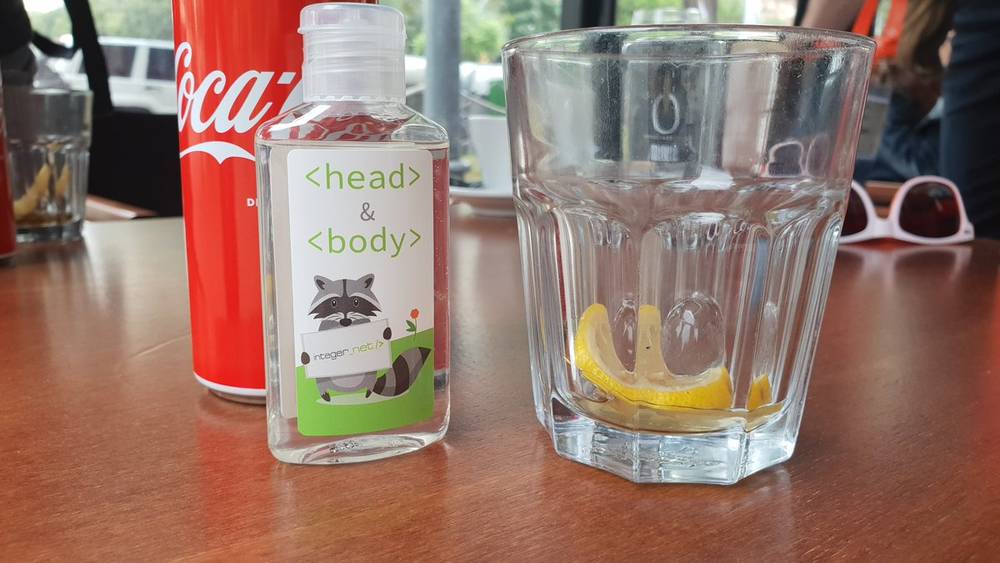 A can of Coca-Cola, a novelty hand sanitizer bottle labeled head and body, and a glass with lemon sit on a table.
