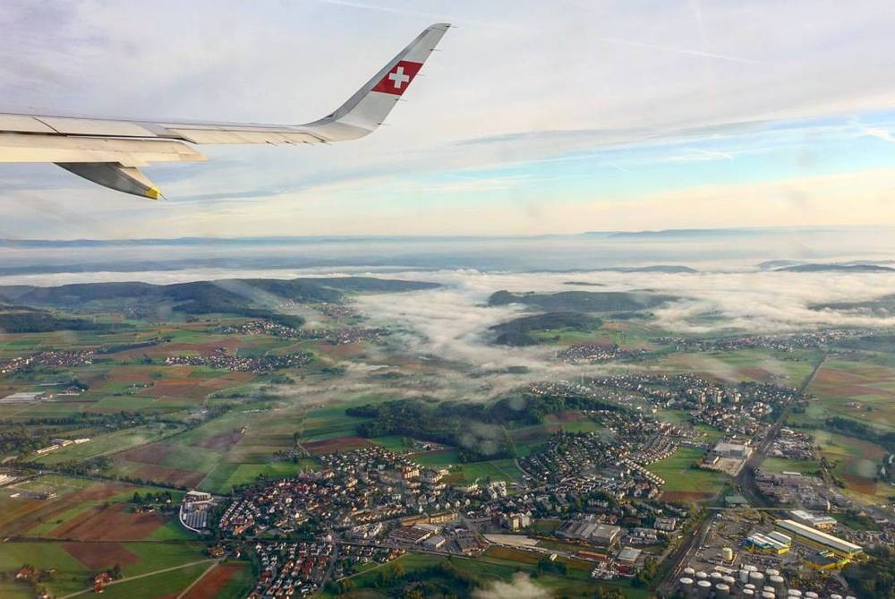 View from an airplane window over a Swiss tail fin, looking down on hills emerging from morning fog.