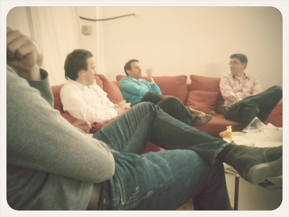 Three men relax with their feet up on a red couch, chatting during a break.