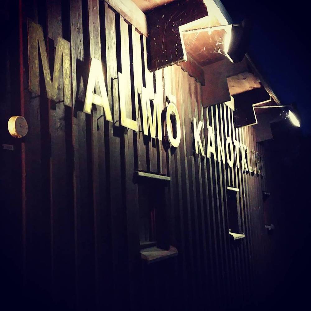 Illuminated letters spelling 'Malmö Kanotklubb' on a wooden building facade at night.