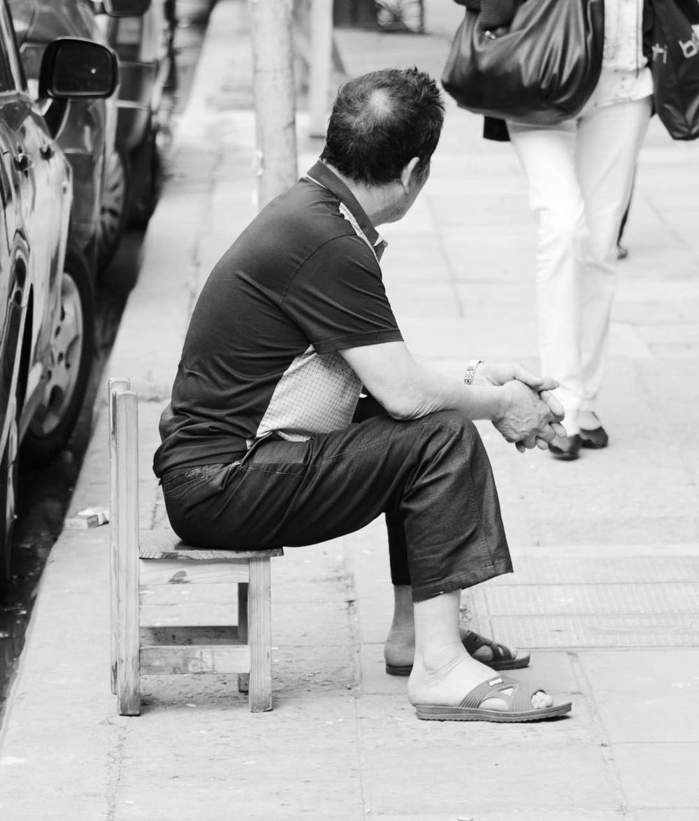A black and white photo of a man sitting on a small stool on a city sidewalk.