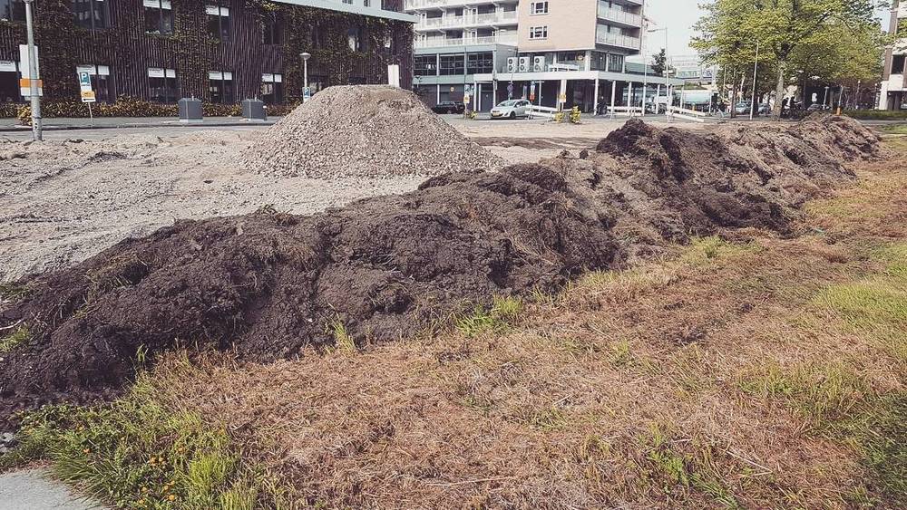 Large piles of excavated dark soil beside a road near modern apartment buildings.