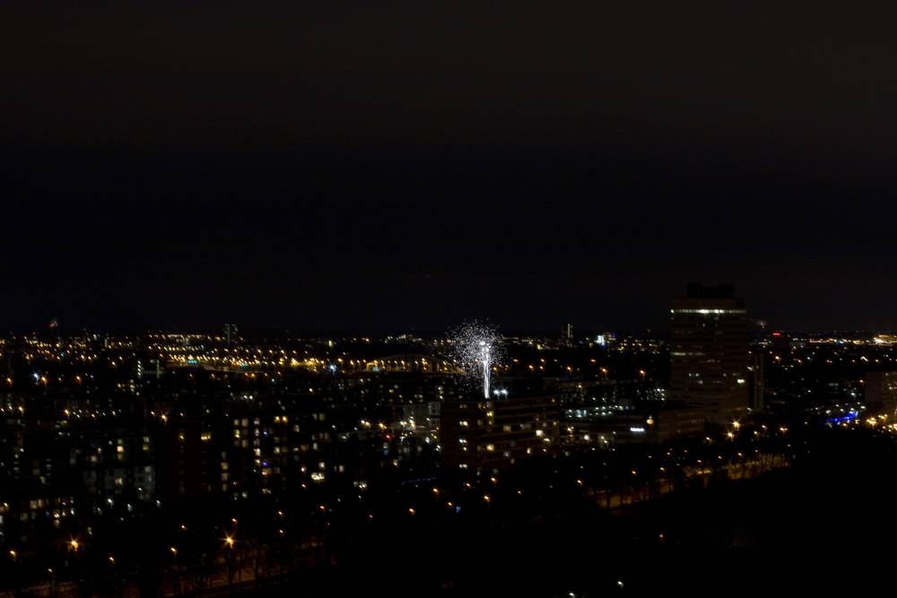 A single firework bursting above a dark city skyline dotted with lights.
