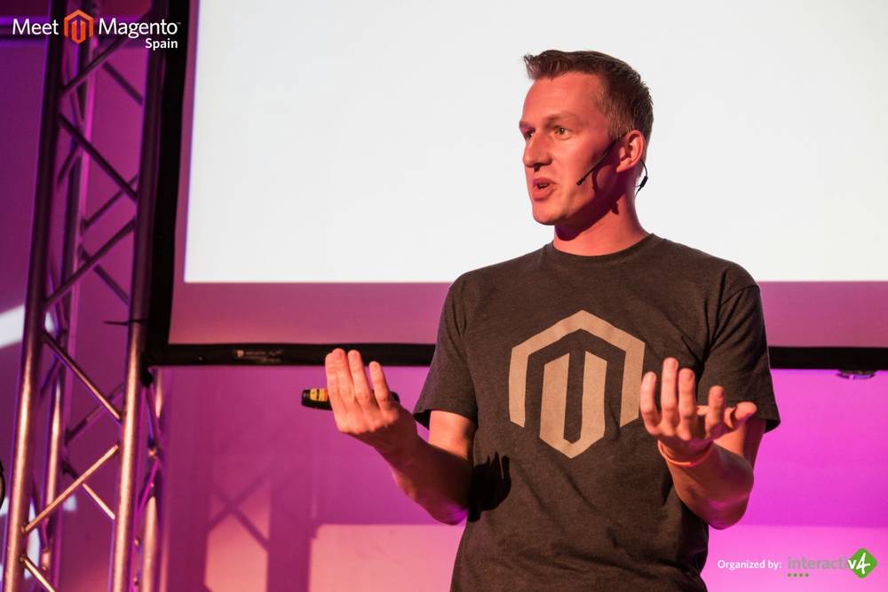 A man in a Magento t-shirt gestures while presenting on a large screen.