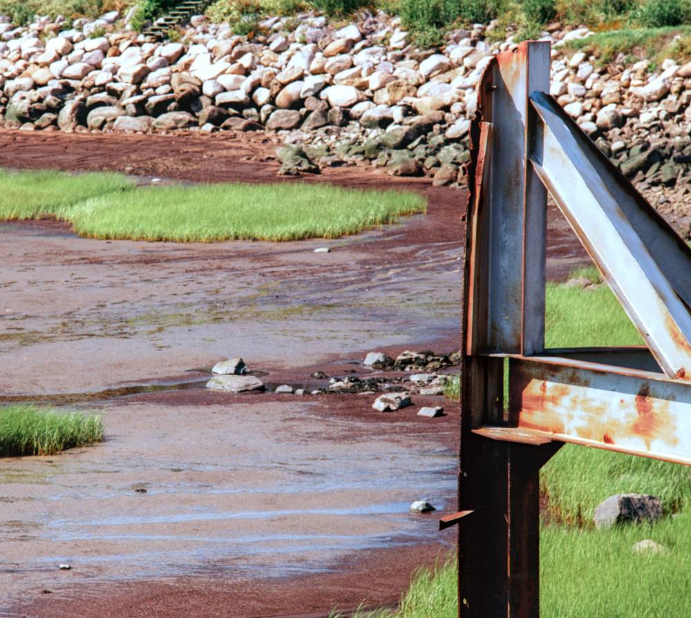 A weathered, rusty metal frame stands on a grassy bank beside a tidal mudflat with patches of green marsh grass.