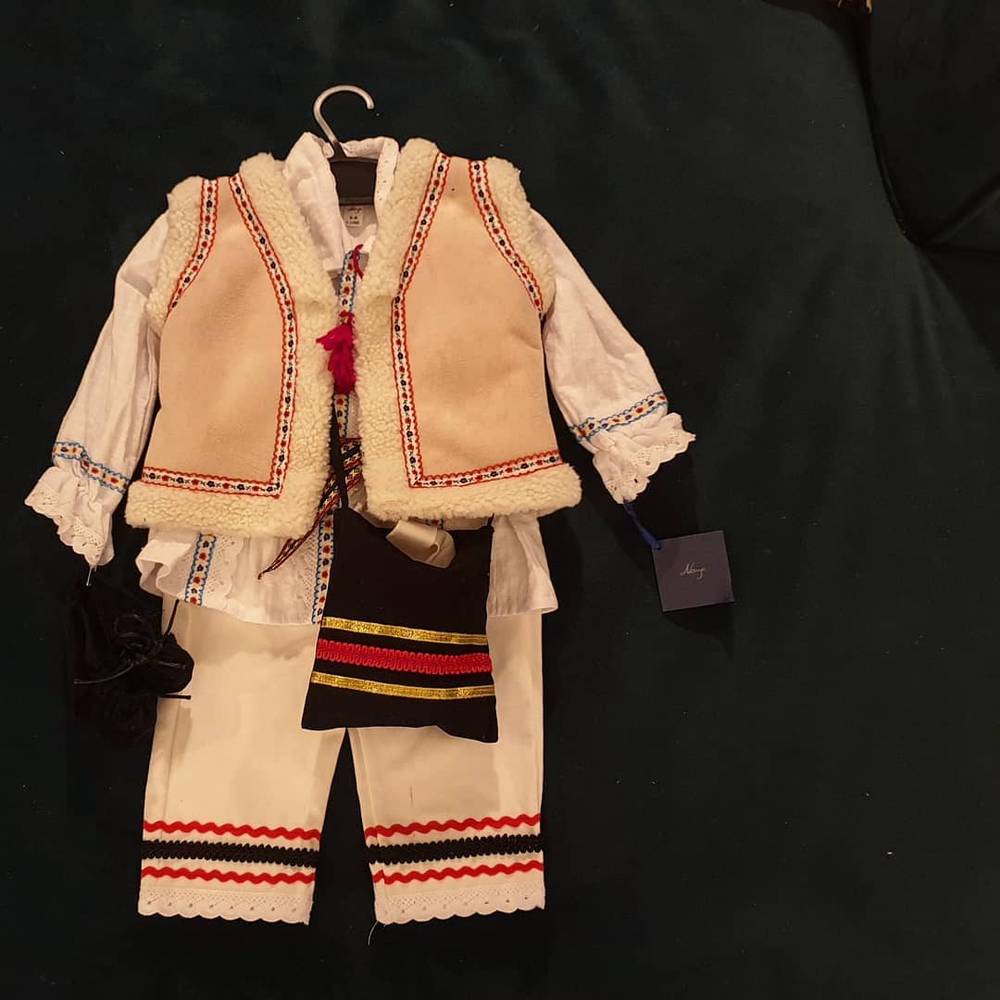 A traditional Romanian folk outfit for a child, with embroidered vest and trim, hangs on a hanger.