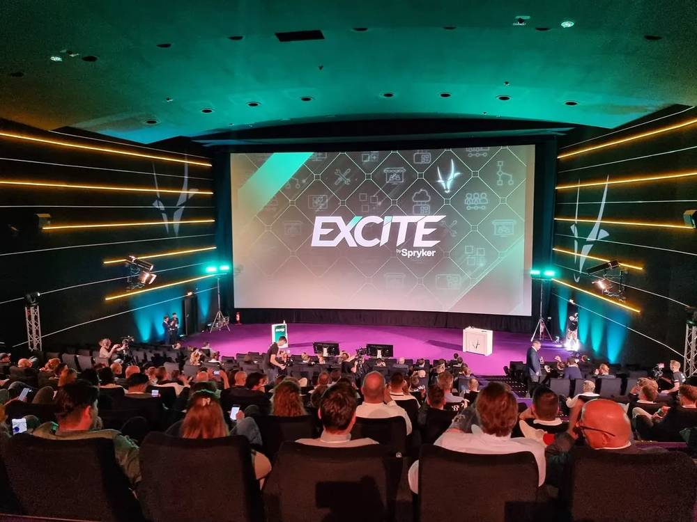 A theater stage with a large screen reading "EXCITE by Spryker," viewed from behind a seated audience.