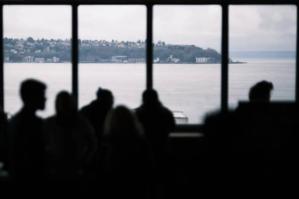 Silhouetted passengers aboard a Washington State Ferry peer through large windows toward the forested hillsides and residential developments of West Seattle across Elliott Bay. The atmospheric haze creates layers of depth, with modern apartment buildings and scattered homes climbing the verdant slopes. The composition frames human contemplation against the Pacific Northwest's signature blend of urban development and natural landscape, captured in soft, overcast light.