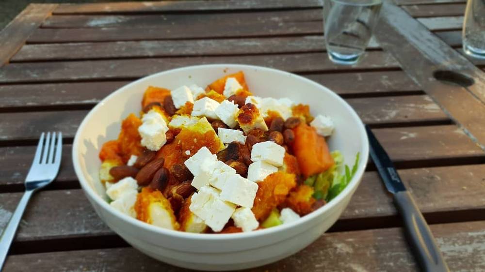 A bowl of roasted pumpkin, feta cheese, and greens on a wooden table.