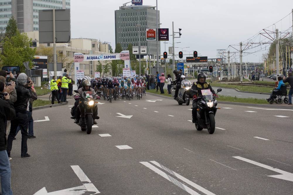 Lead motorbikes precede the approaching peloton as spectators and photographers watch.