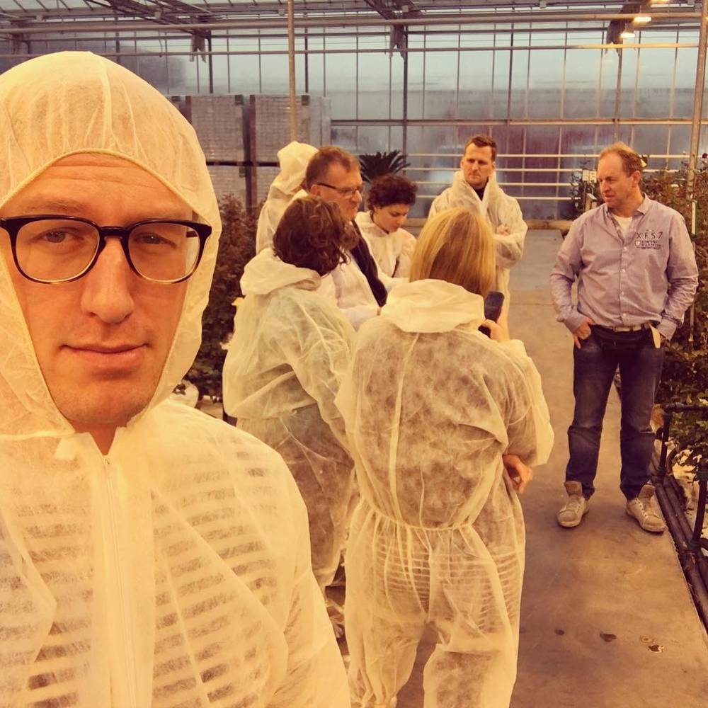 A man in glasses wearing a white disposable coverall, with a group of people dressed the same way in a greenhouse behind him.