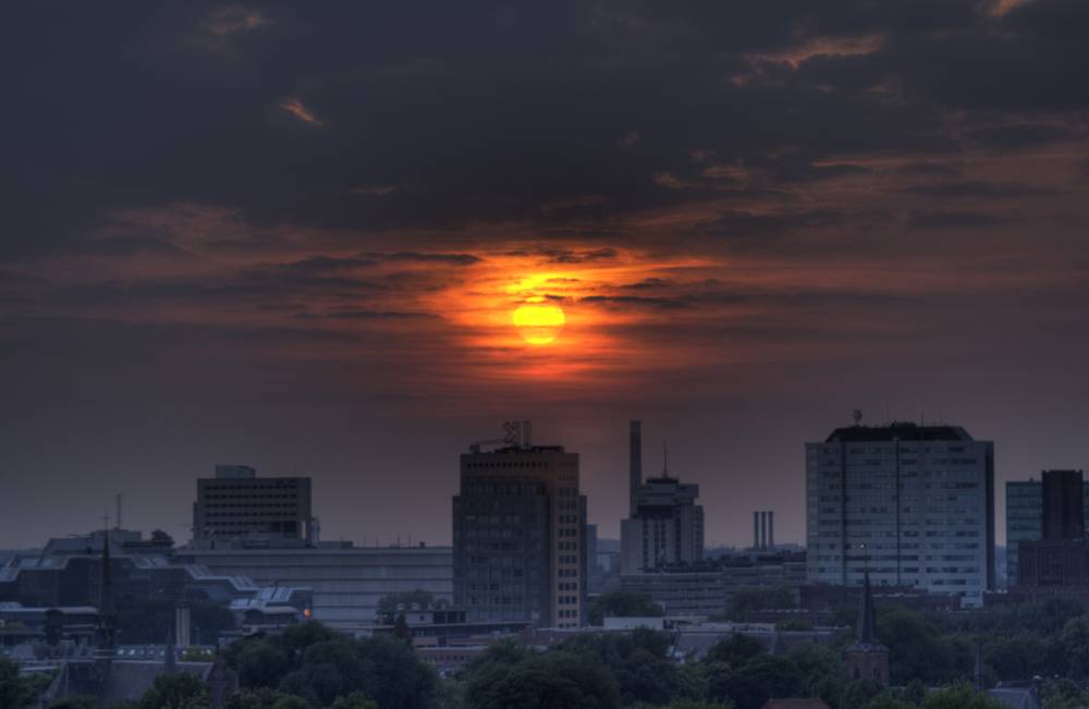 The sun glows orange through layered clouds at sunset over a city skyline.