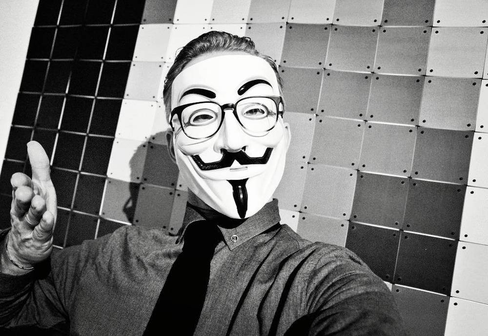 A black and white selfie of a man wearing a Guy Fawkes mask against a tiled wall.