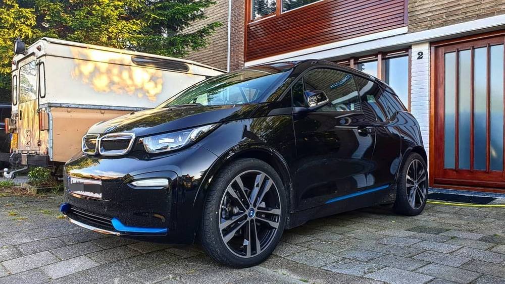 A black BMW i3 electric car is parked outside a building decorated with lights at dusk.