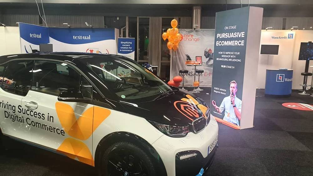 A branded BMW i3 car sits at an event booth beneath a "Driving Success in Digital Commerce" banner, a man presenting behind it.