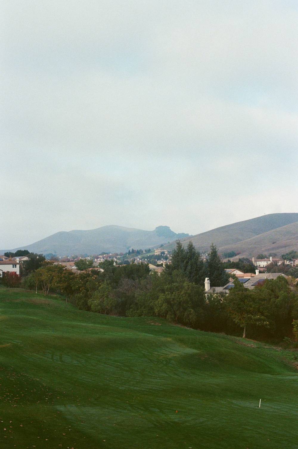 a sprawling green lawn of a golf course surrounded by trees and houses with mountainy hills in the background.