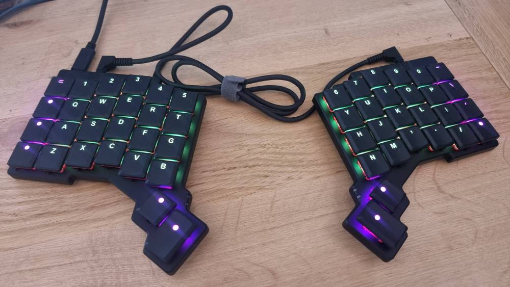A split ergonomic keyboard with colourful backlit keys on a wooden desk.