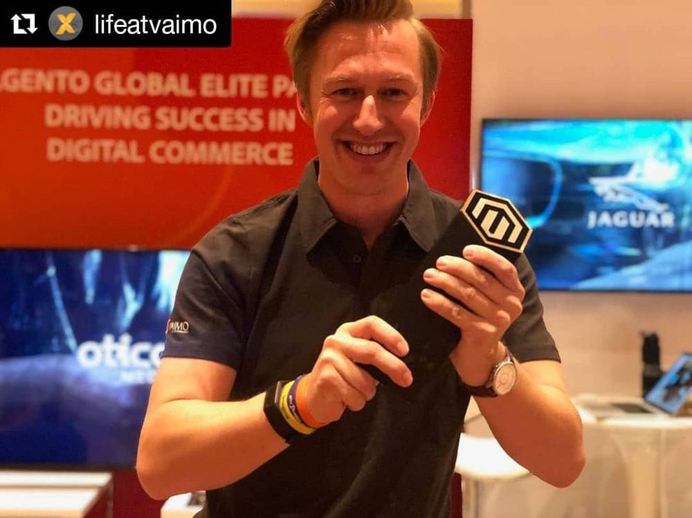 A man holds a hexagon-shaped Magento badge while smiling at an event booth.