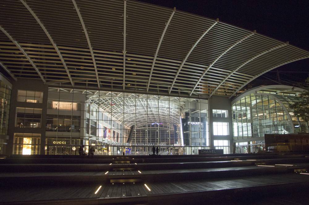 A curved glass canopy building glows above an empty boardwalk lined with light strips at night.
