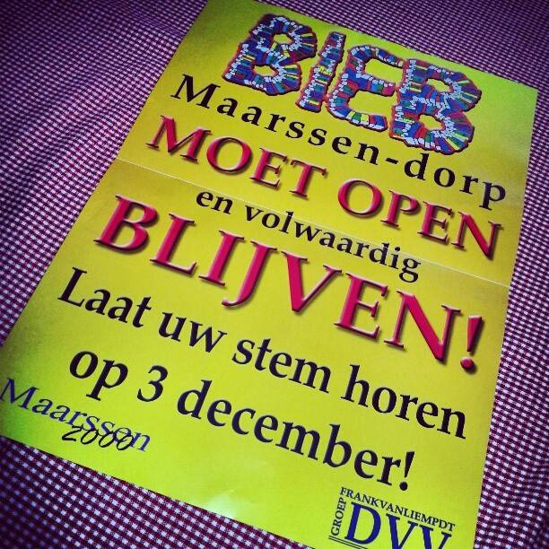 A yellow flyer reading 'Bieb Maarssen-dorp moet open en volwaardig blijven! Laat uw stem horen op 3 december!' lying on a striped doormat.