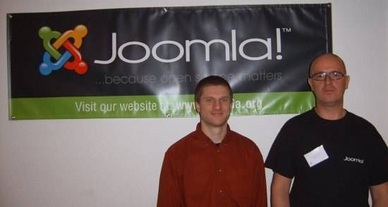 Two men pose together in front of a Joomla banner at a conference.