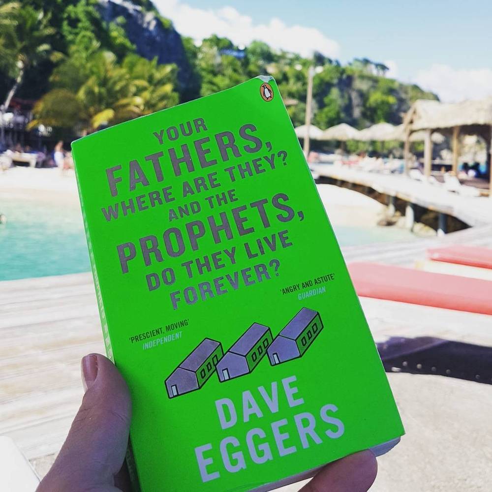 A hand holding the book "Your Fathers, Where Are They? And the Prophets, Do They Live Forever?" by Dave Eggers on a beach.