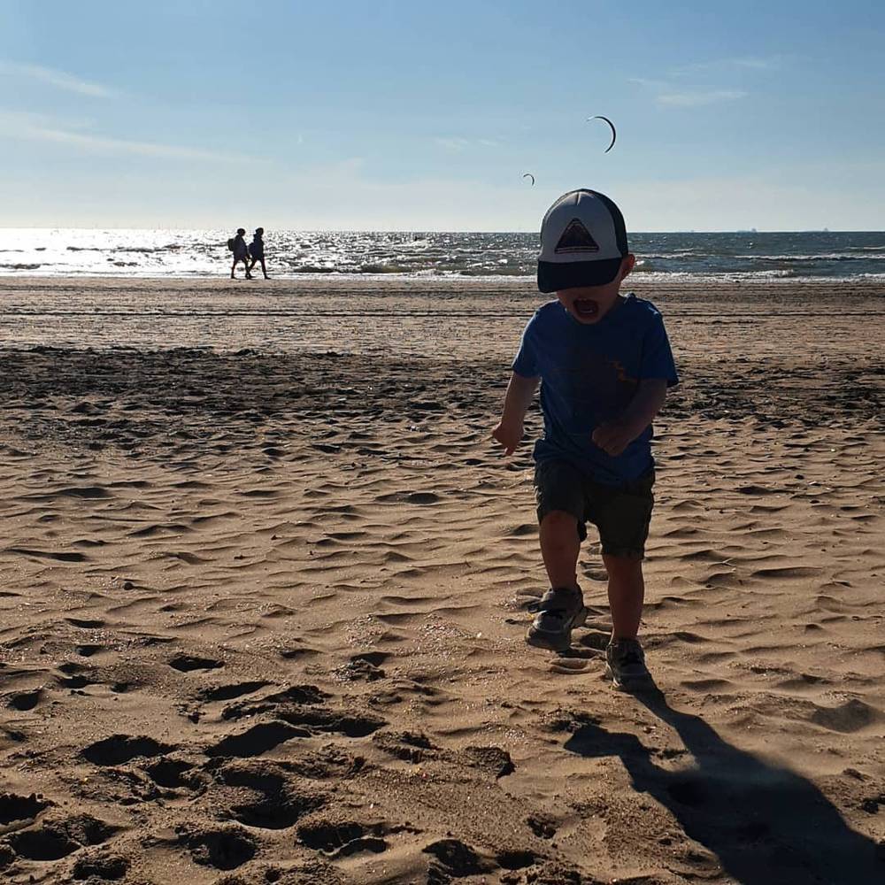 A child in a cap walking across a sandy beach, with horse riders and a kite visible in the distance.