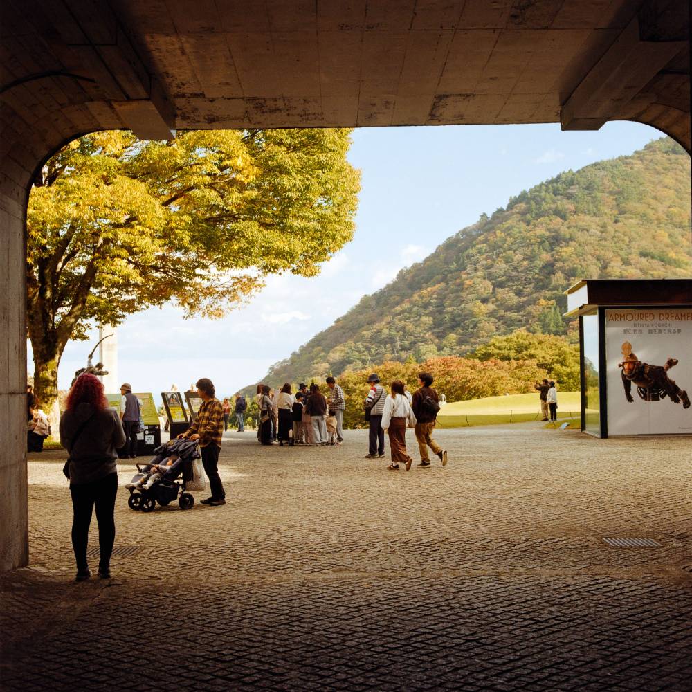 A view looking through the end of a rounded rectangular tunnel. Outside the tunnel, people miss about under a tree
