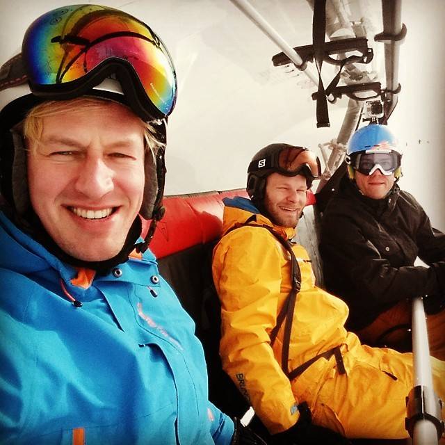 Three people ride a chairlift together, smiling, wearing ski helmets and goggles.