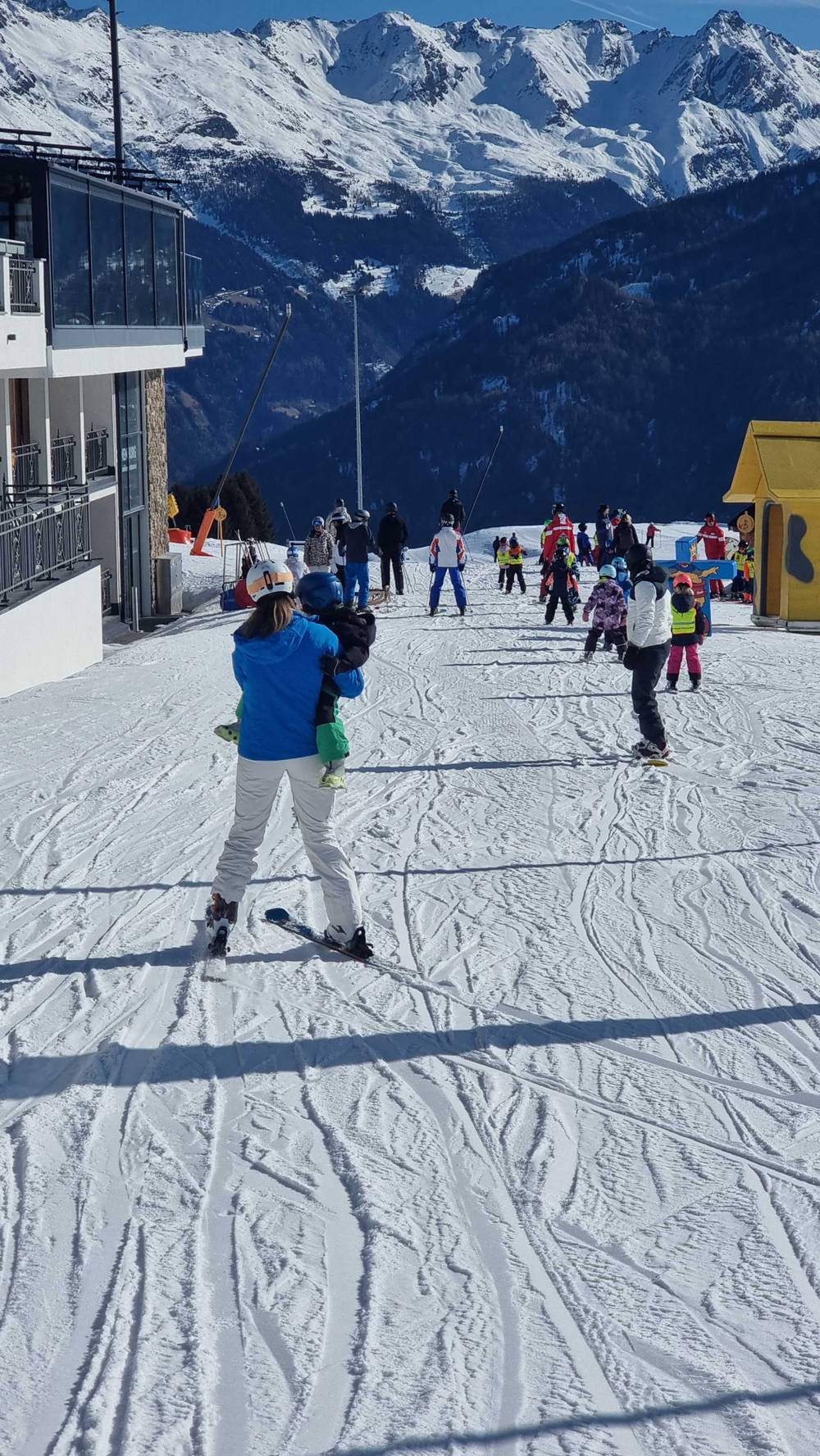 Two skiers make their way down a slope near mountain lodge buildings.